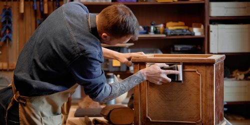 Man crafting intricate antique furniture piece in woodworking workshop surrounded by tools and materials, meticulously sanding the wood surface